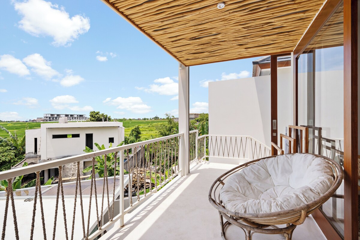 A balcony with a wicker chair, a view of a building, and a railing.