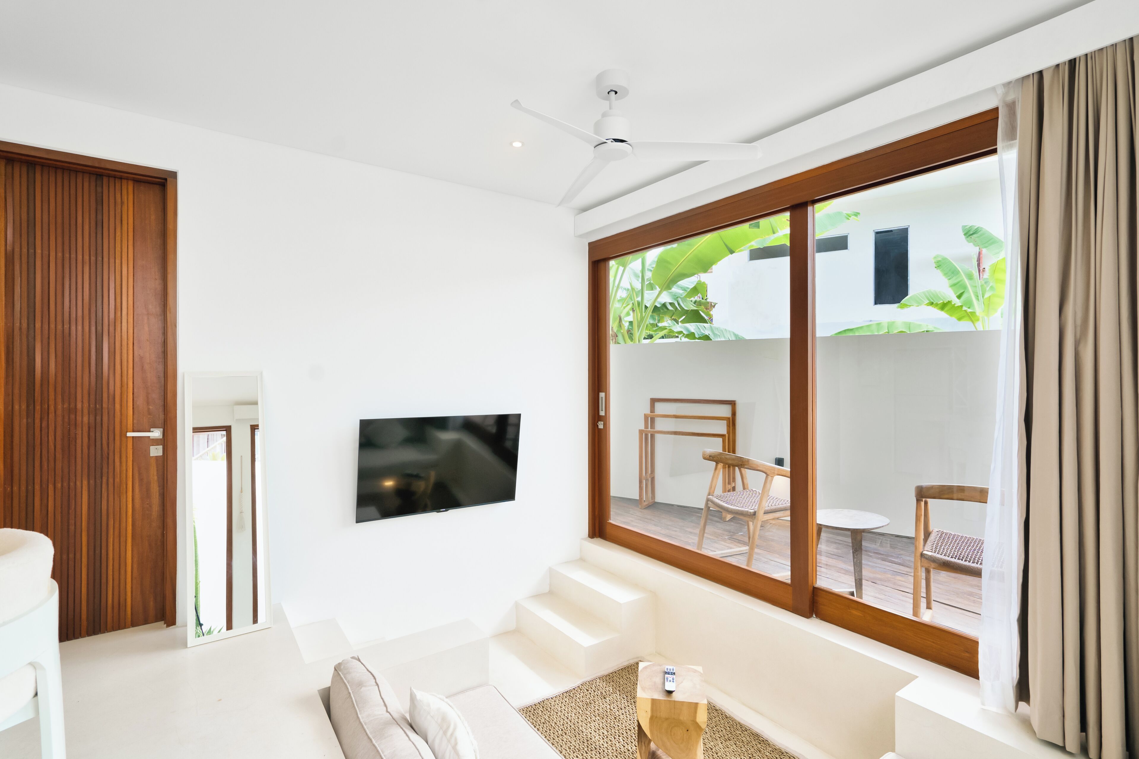 A modern living room with a flat-screen TV, a sofa, and a coffee table. There is a sliding glass door leading to an outdoor area with a table and chairs.