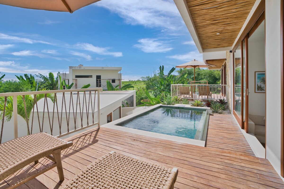 A wooden deck with a pool, outdoor furniture, and a view of greenery and buildings.