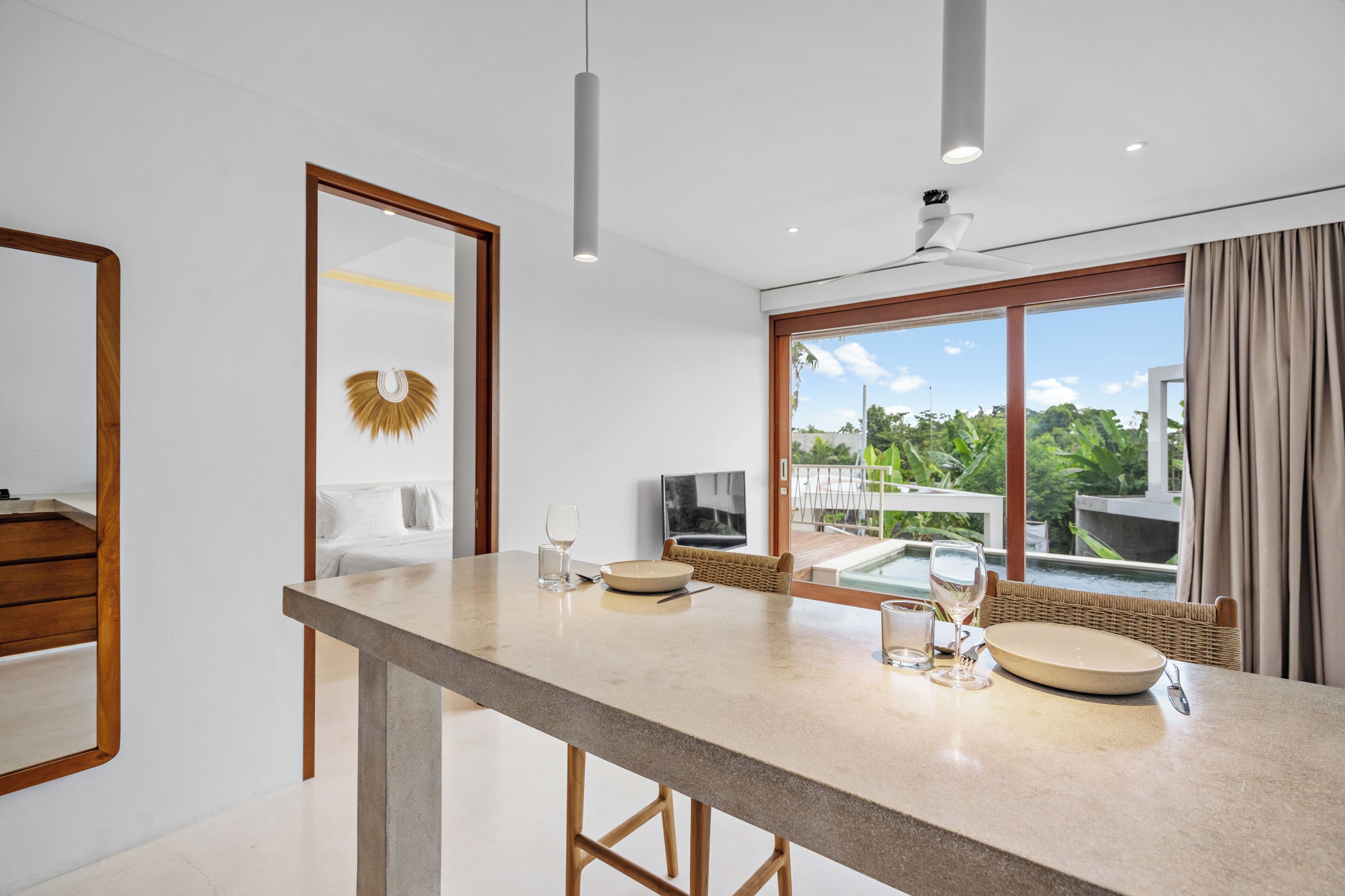 A modern dining area with a long table, chairs, and a view of a pool and greenery outside.