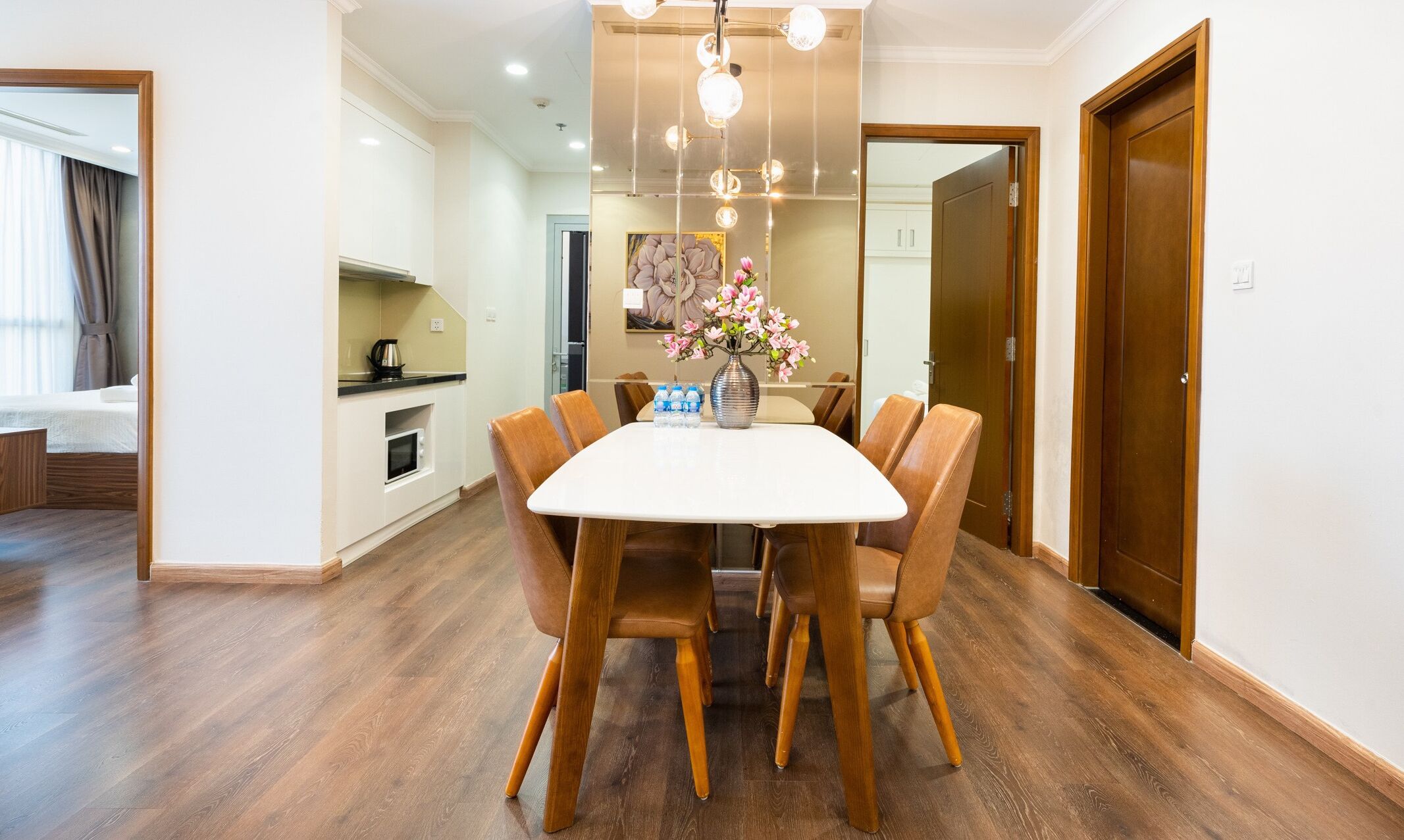 Un comedor con una mesa blanca, seis sillas de madera, un florero con flores y una cocina al fondo.