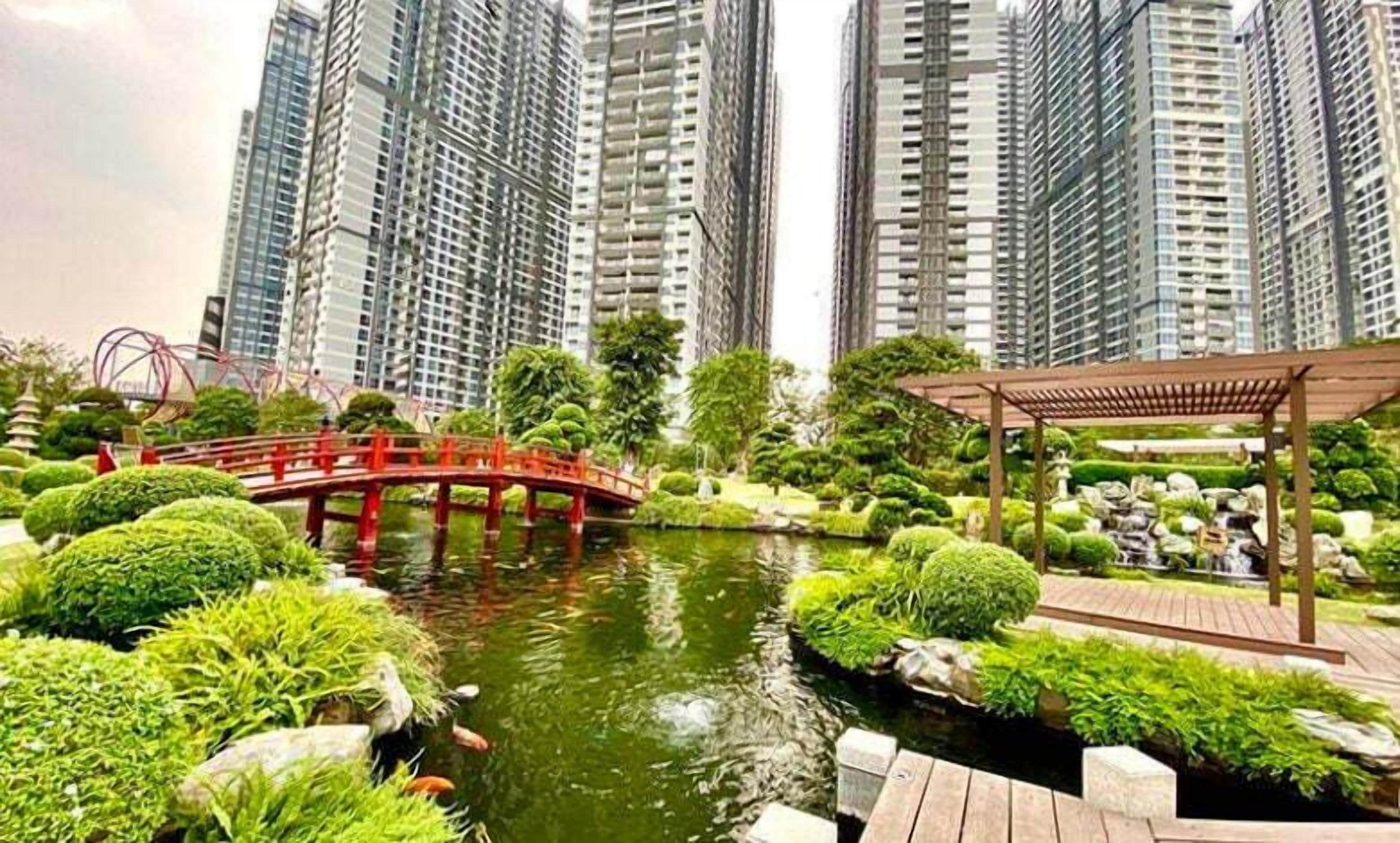 A serene park with a pond, surrounded by lush greenery and a red bridge. Tall residential buildings are in the background.