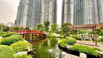 A serene park with a pond, surrounded by lush greenery and a red bridge. Tall residential buildings are in the background.
