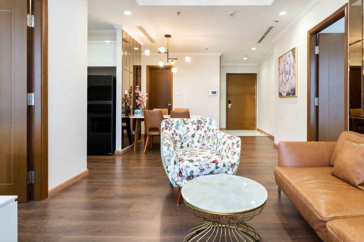 A hotel lobby with a wooden floor, a round marble table, a floral armchair, a leather sofa, and a dining area in the background.