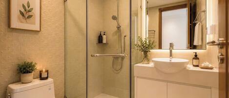 A bathroom with a glass-enclosed shower, a white toilet, a sink with a mirror, and a framed plant on the wall.
