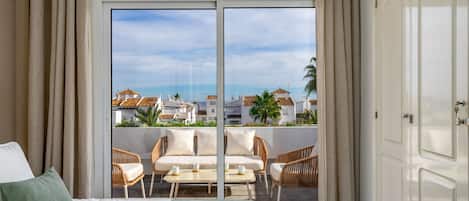 Quarto de hotel com varanda, uma cama, mobiliário de vime e vista para edifícios e palmeiras.