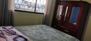 Ein Schlafzimmer mit Bett, Schrank, einem Fenster mit Blick auf Gebäude und Vorhängen.