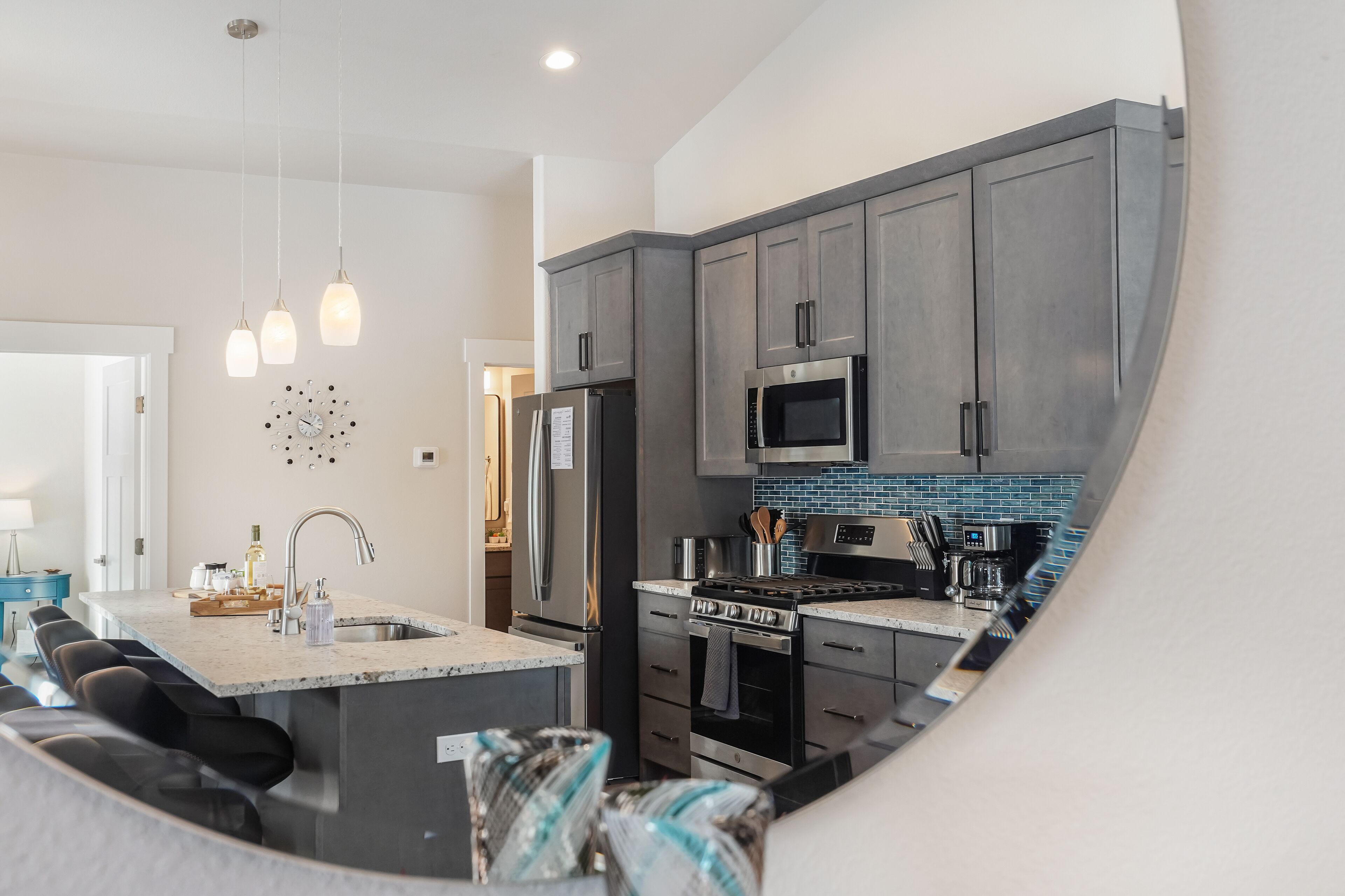 A modern kitchen with grey cabinets, a stainless steel refrigerator, a microwave, and a built-in oven. There is a large island with a sink and seating.