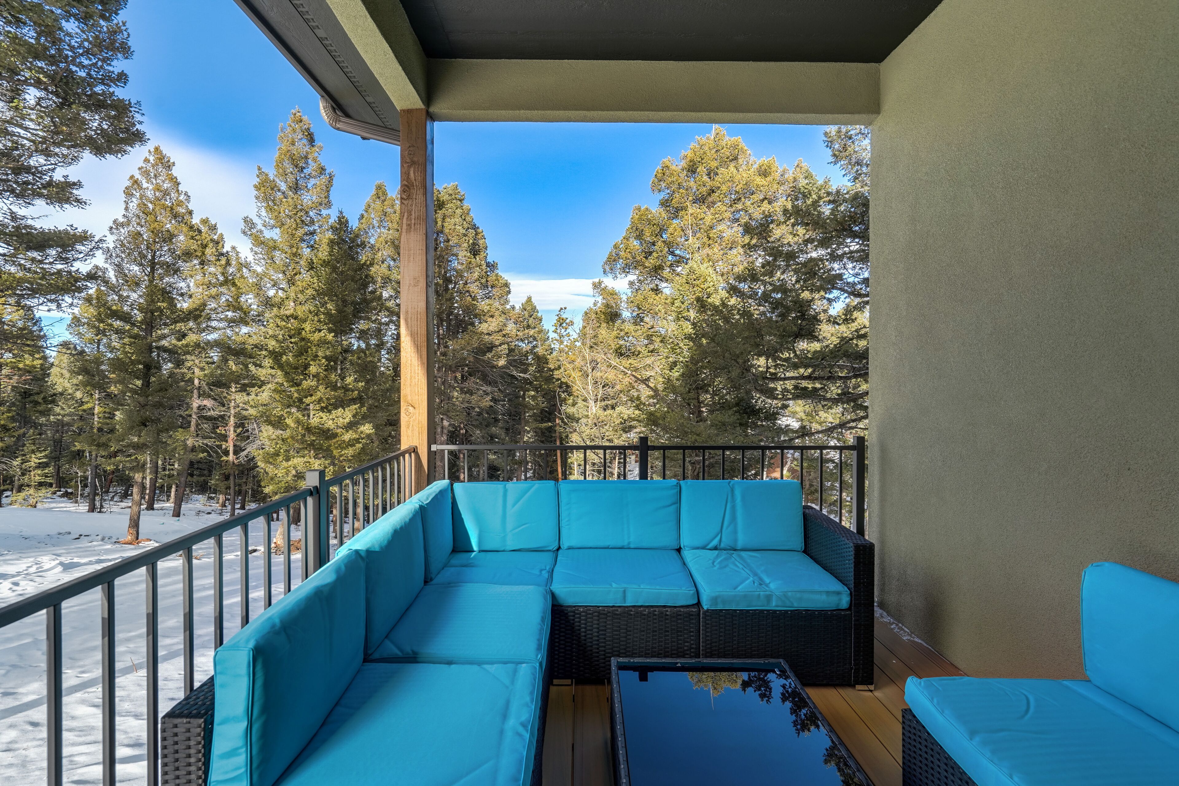 Un espace extérieur aménagé, abrité et doté d’un canapé bleu rembourré et d’une petite table, le tout entouré d’une rambarde en métal, offrant une vue sur un paysage enneigé et de grands arbres.