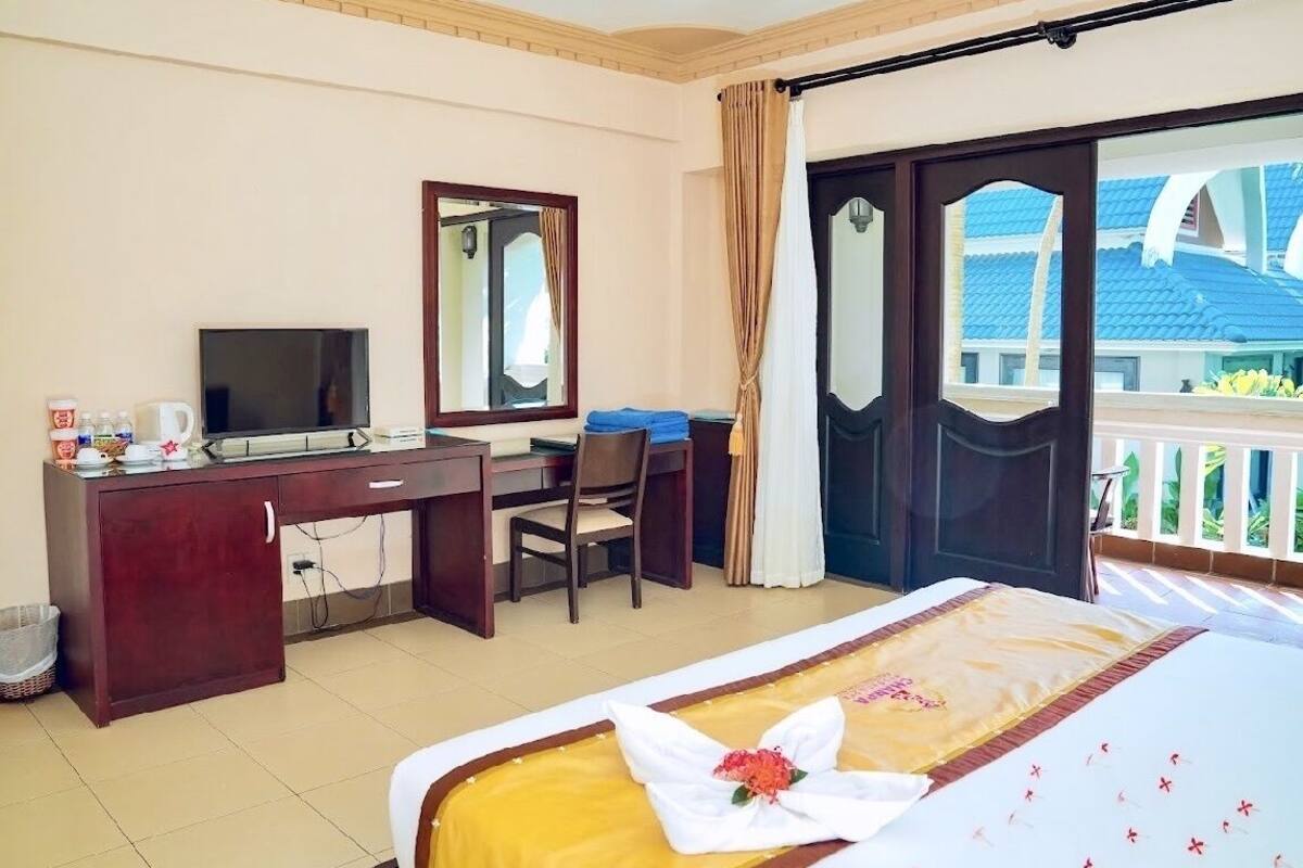 Una habitación de hotel con cama, escritorio, televisión y balcón con vista a un edificio de techos azules.