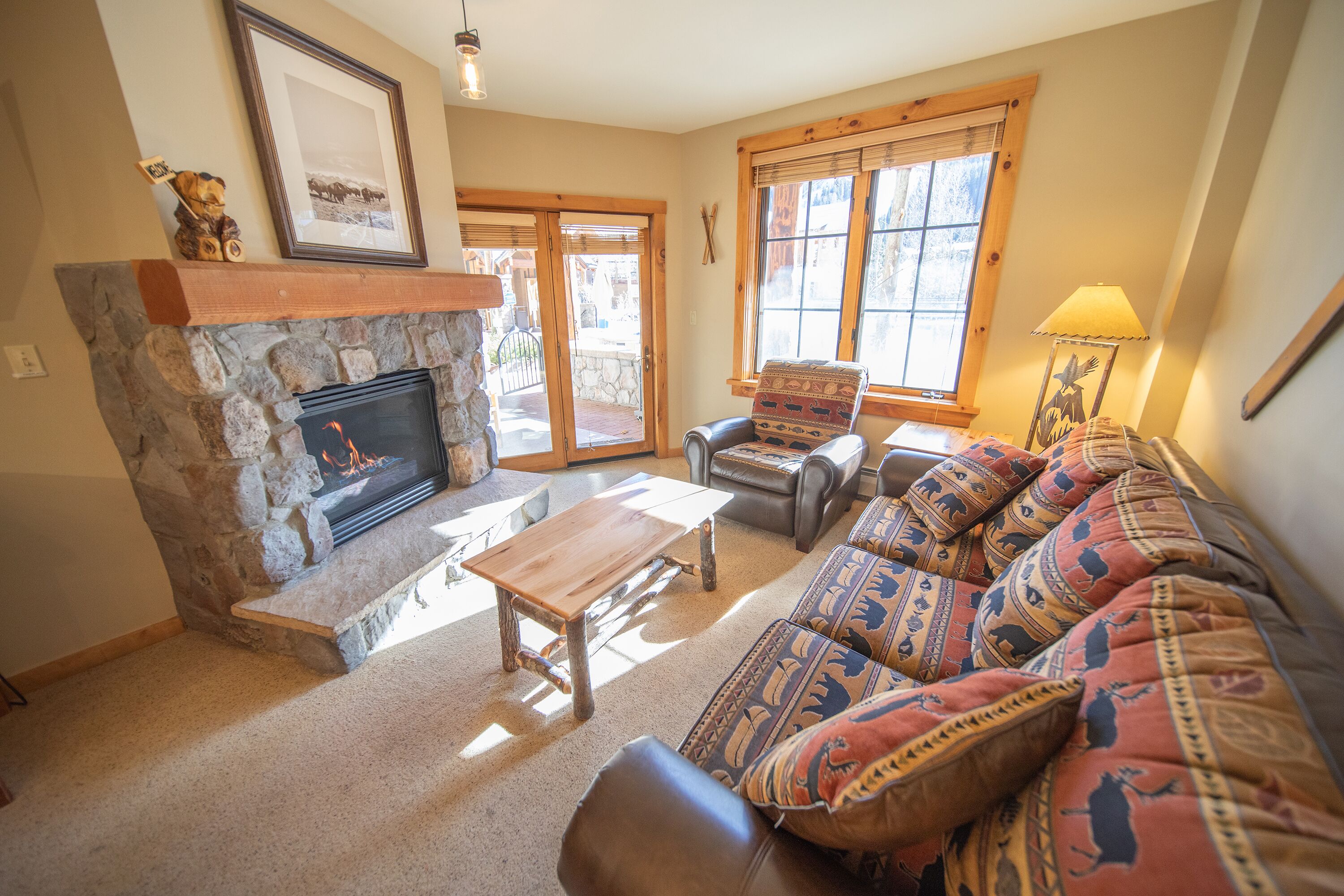 A cozy living room with a stone fireplace, a sofa with patterned cushions, a wooden coffee table, and a framed picture on the wall.