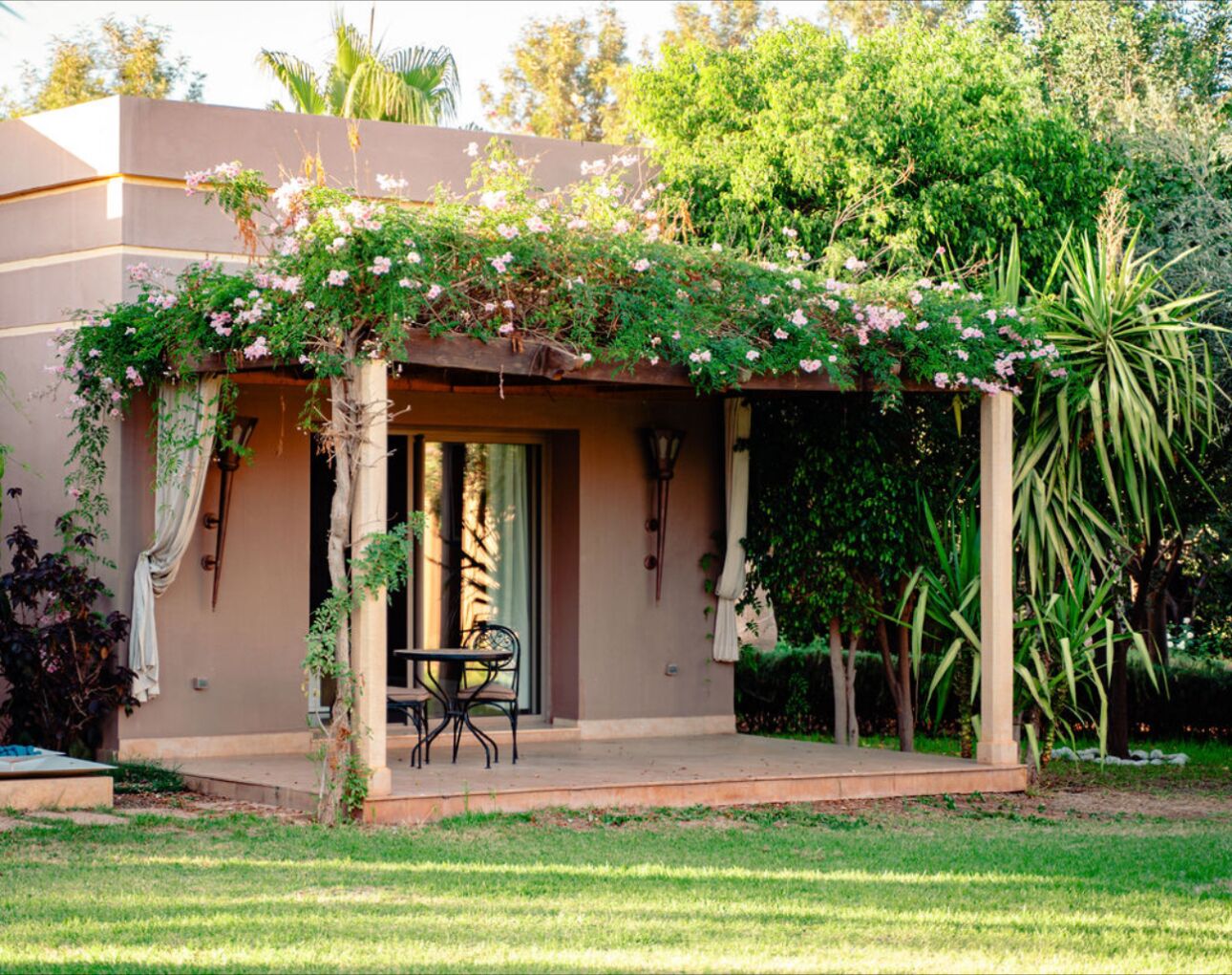 A covered outdoor seating area with a table and chairs, surrounded by lush greenery and flowering plants.