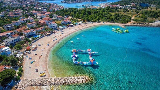 Vue aérienne d’une ville côtière avec une plage dégagée, un port de plaisance rempli de bateaux et des immeubles résidentiels.