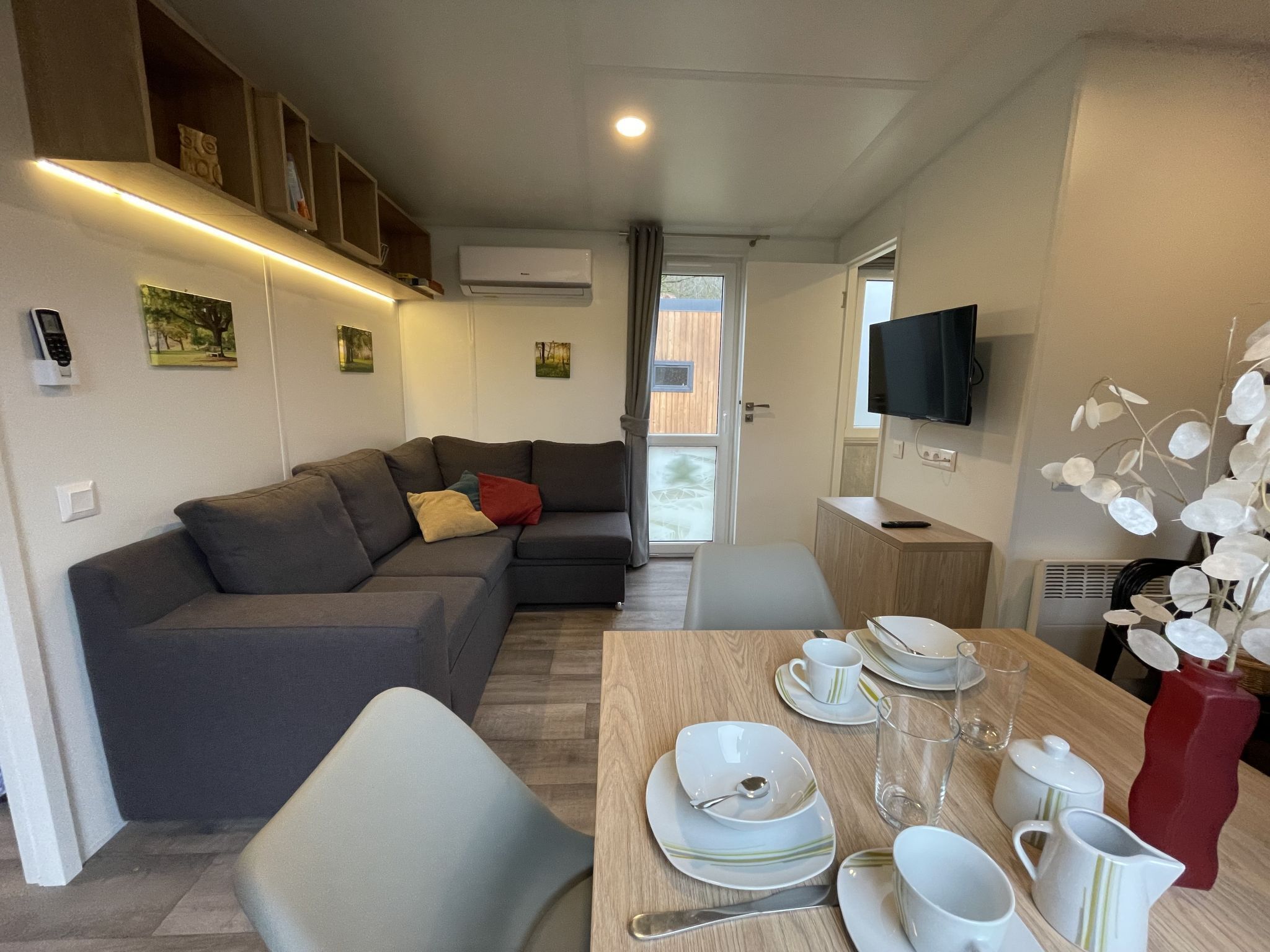 Ein modernes Wohnzimmer mit einem grauen Ecksofa, einem Esstisch mit weißen Tellern, Tassen und einer Karaffe, und einem an der Wand befestigten Fernseher.