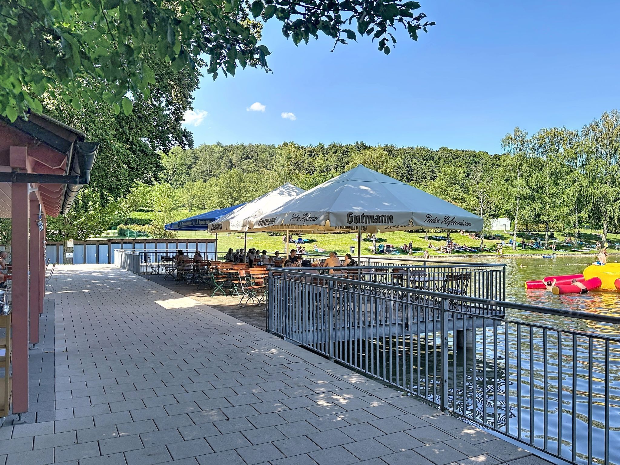 Ein Außenbereich am Flussufer mit Tischen und Stühlen, einem überdachten Pavillon und einer Metallreling entlang des Wassers.