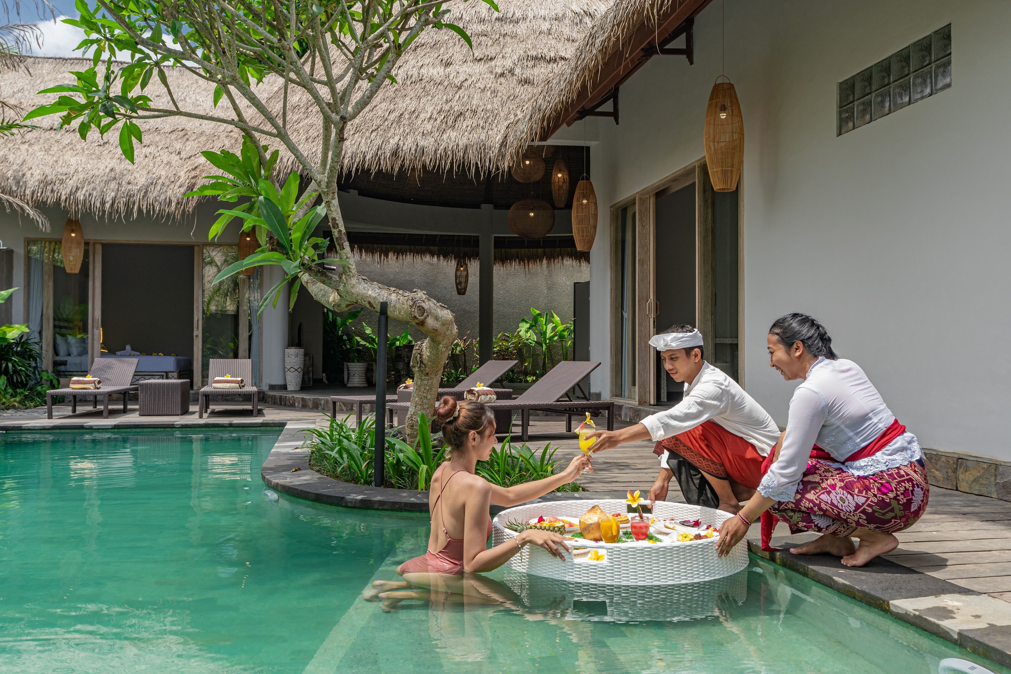 A poolside dining area with a round table, lounge chairs, and a thatched roof structure.