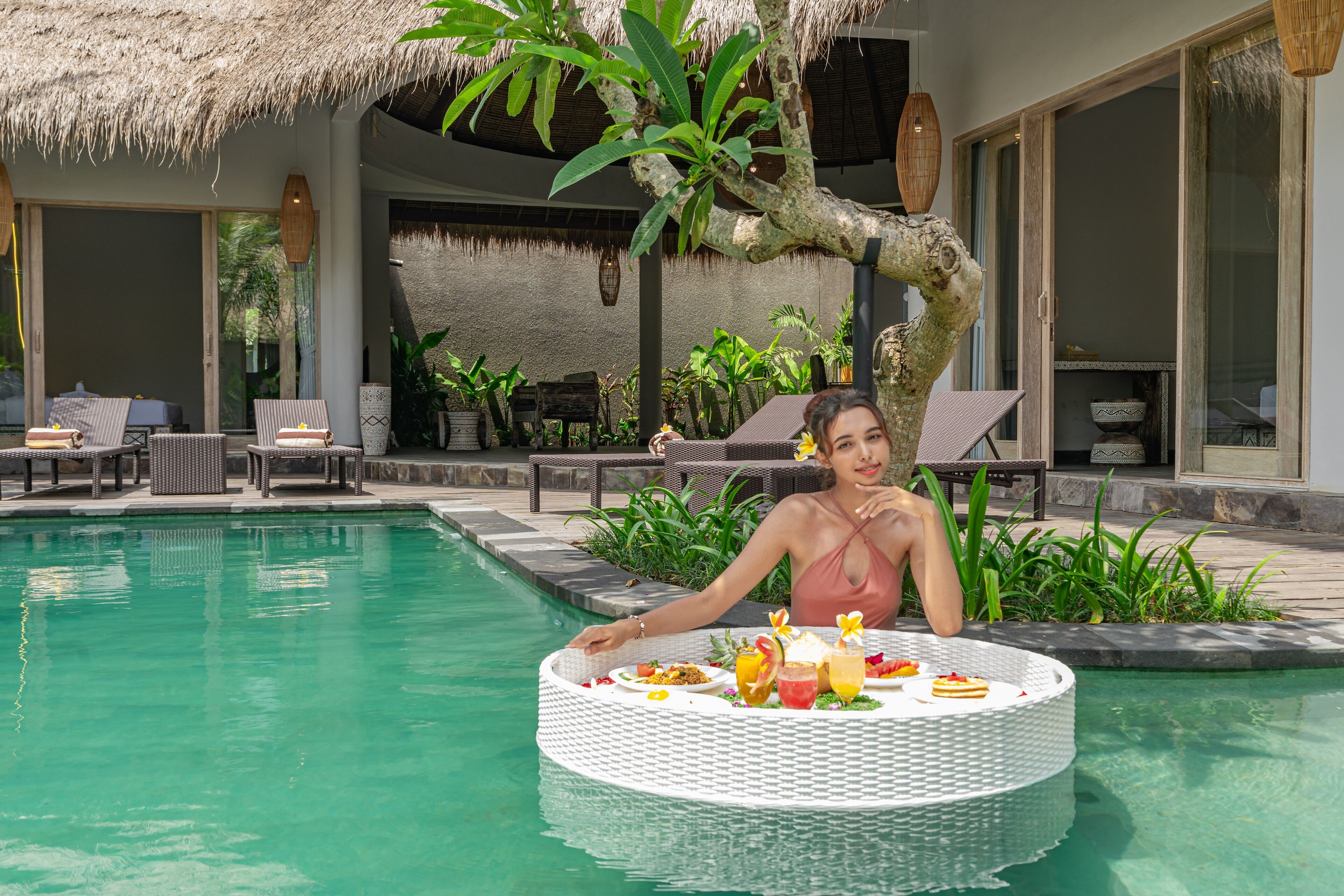 A poolside dining area with a round table, lounge chairs, and a thatched roof structure.