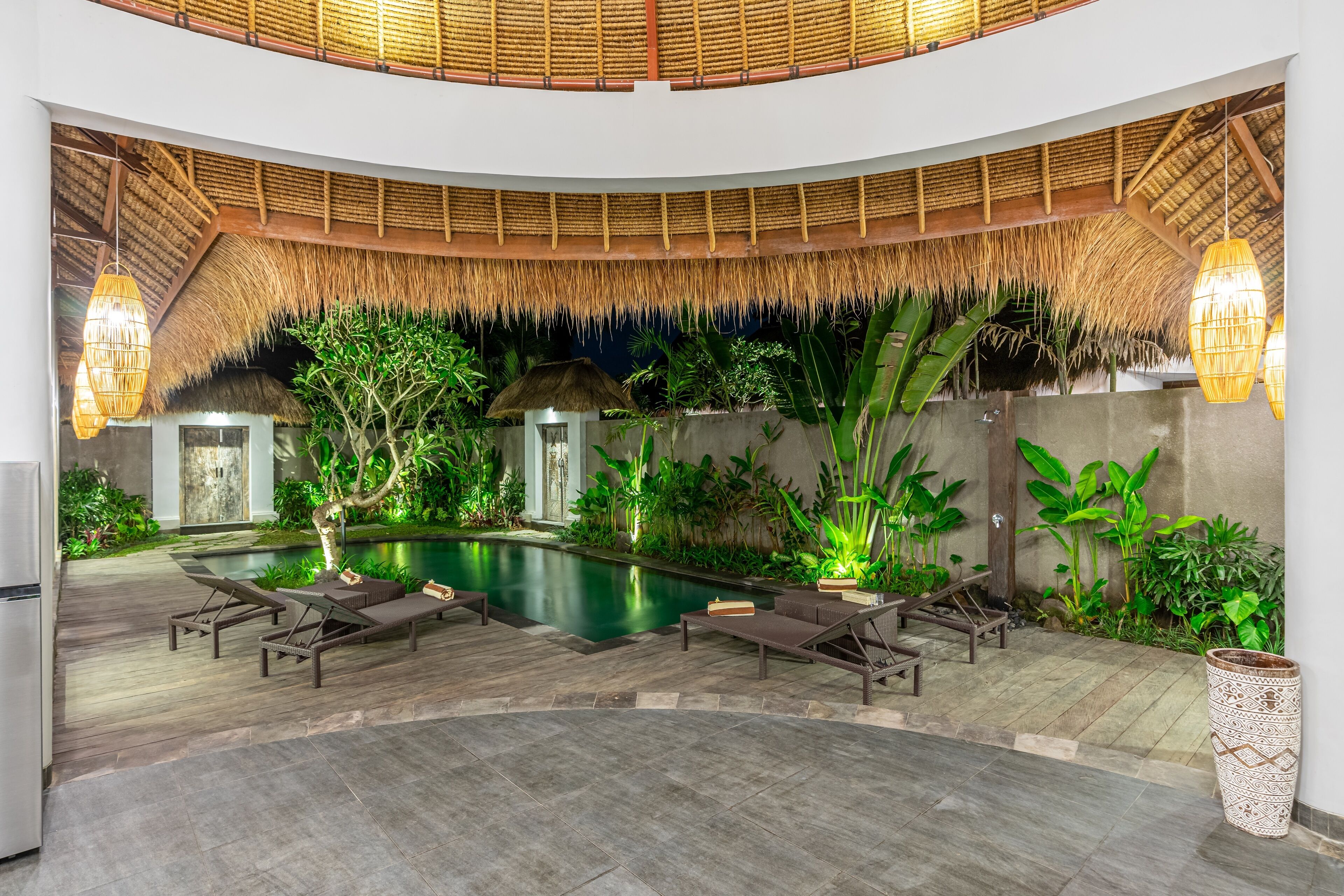 A circular pool area with lounge chairs, a thatched roof, and lush greenery.