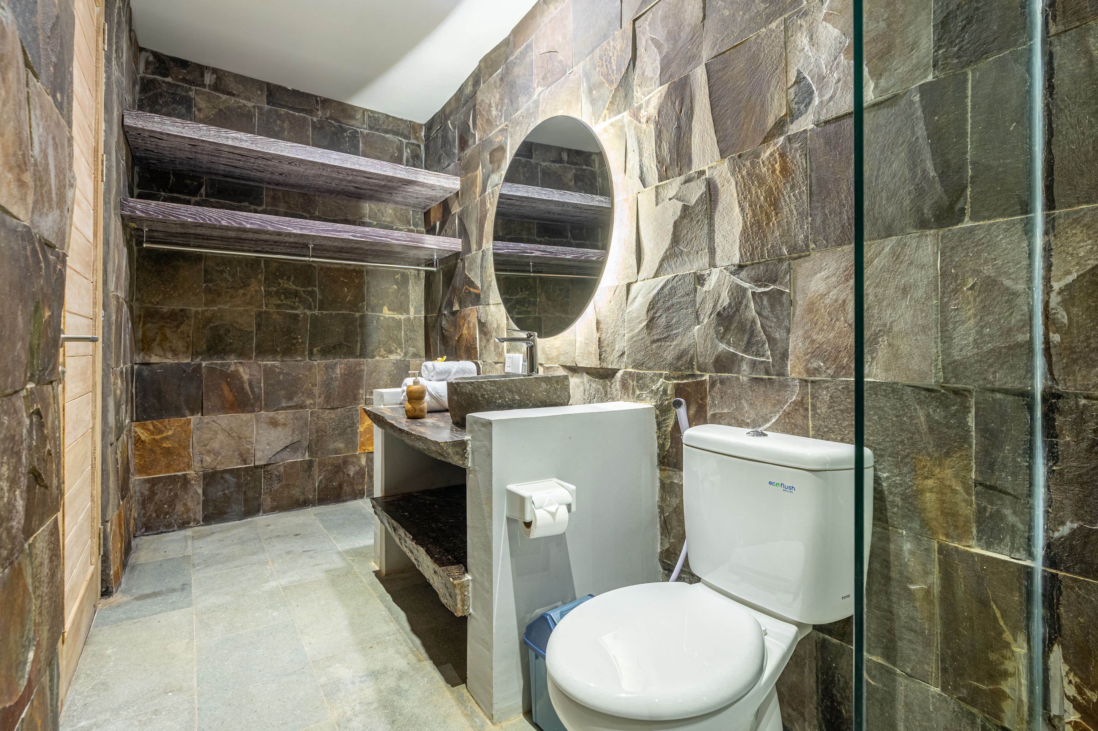 Un baño con pared de piedra, repisa de madera, inodoro y espejo.