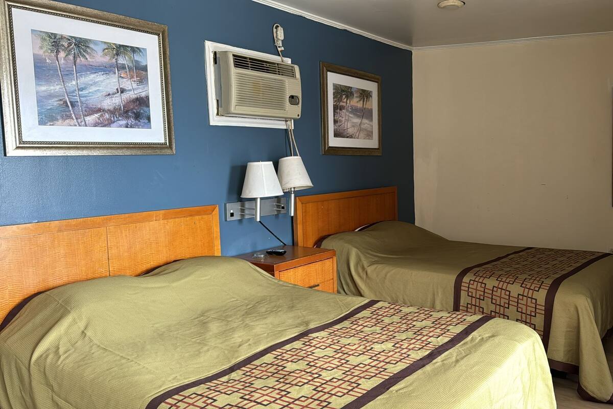 A hotel room with two beds, a nightstand with a lamp, a wall-mounted air conditioner, and two framed pictures of a beach scene.