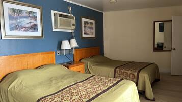 A hotel room with two beds, a nightstand with a lamp, a wall-mounted air conditioner, and two framed pictures of a beach scene.
