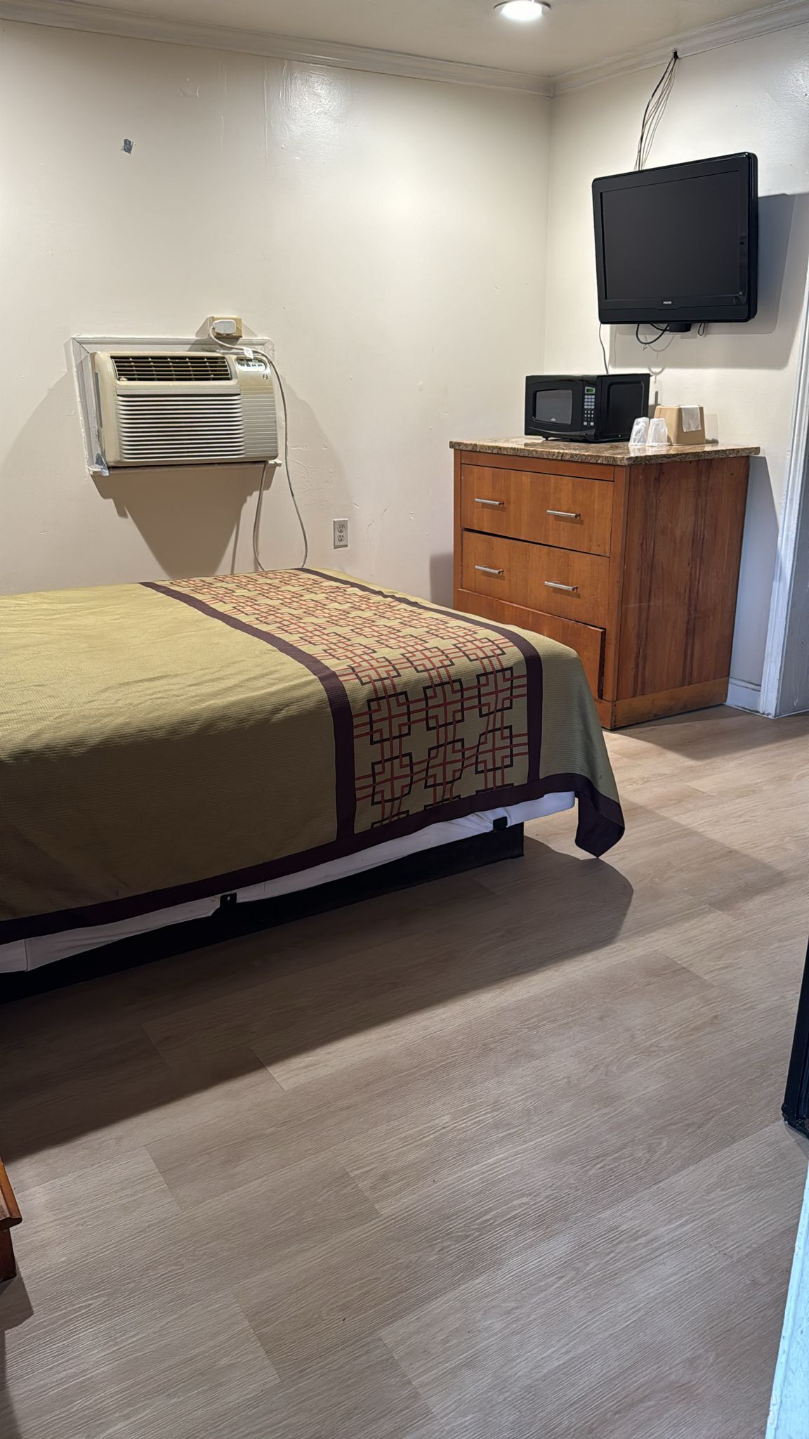 A bedroom with a bed, a wooden dresser, a wall-mounted TV, and an air conditioning unit.