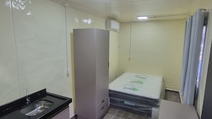 A compact hotel room with a kitchenette, a bed with a mattress and pillows, a wardrobe, and a small table with a stool. - Pousada Vila Verde Containers (Barra Velha)