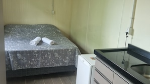 A small, compact room with a bed, a small white refrigerator, a sink, and a window. - Pousada Vila Verde Containers (Barra Velha)