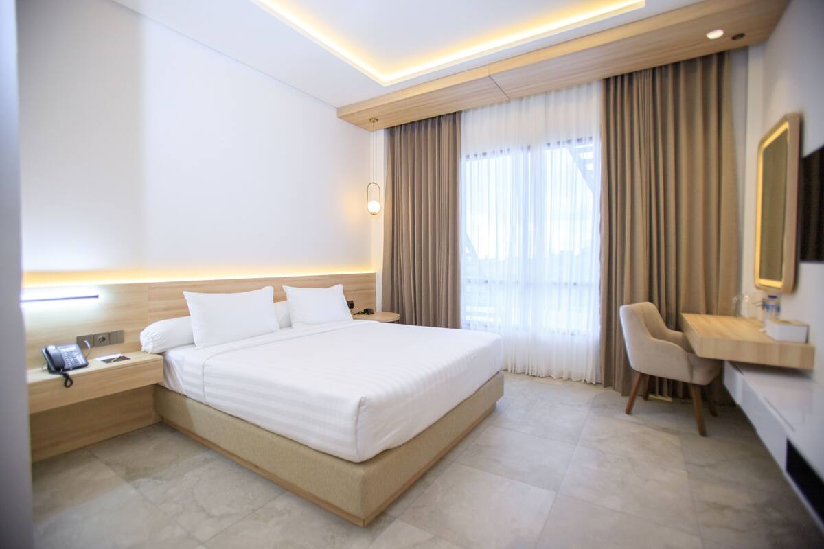 Una habitación de hotel moderna con una cama grande, una mesita de noche con teléfono, un escritorio con silla y un espejo amplio.