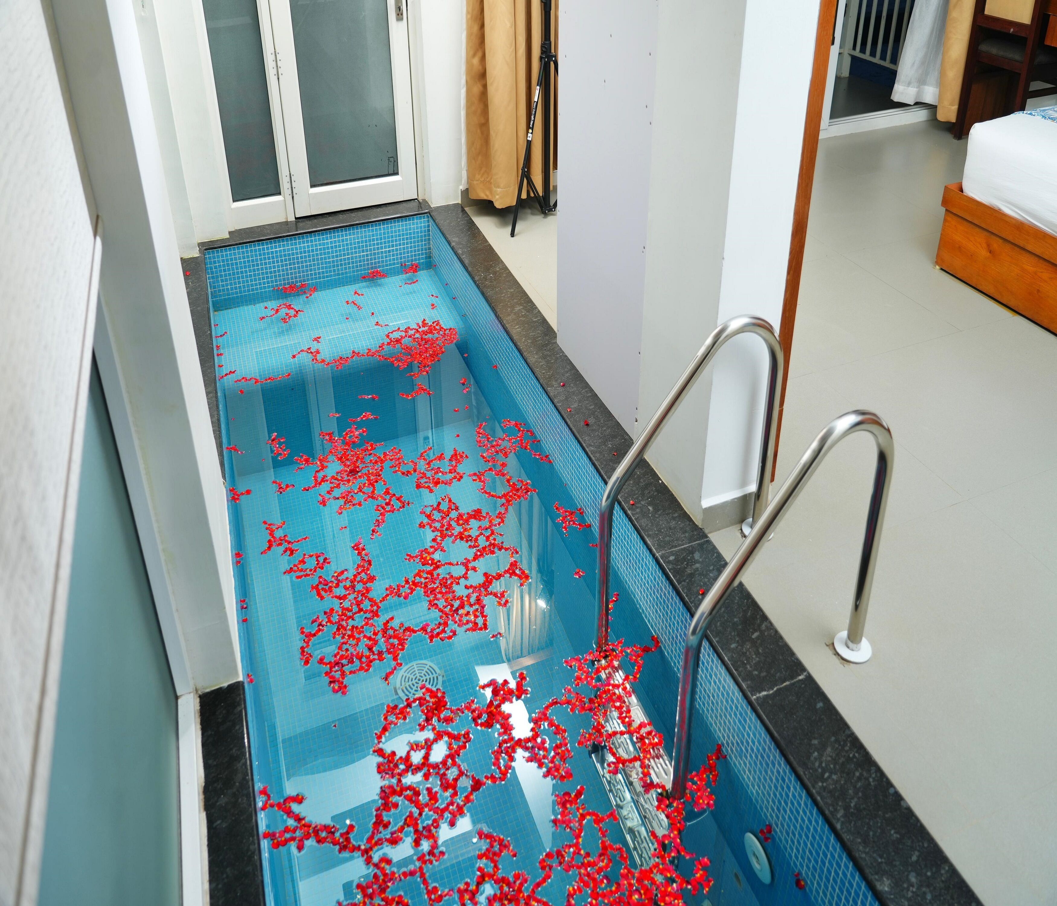 A swimming pool with red flower petals scattered on the water, a metal ladder, and a bed in the background.