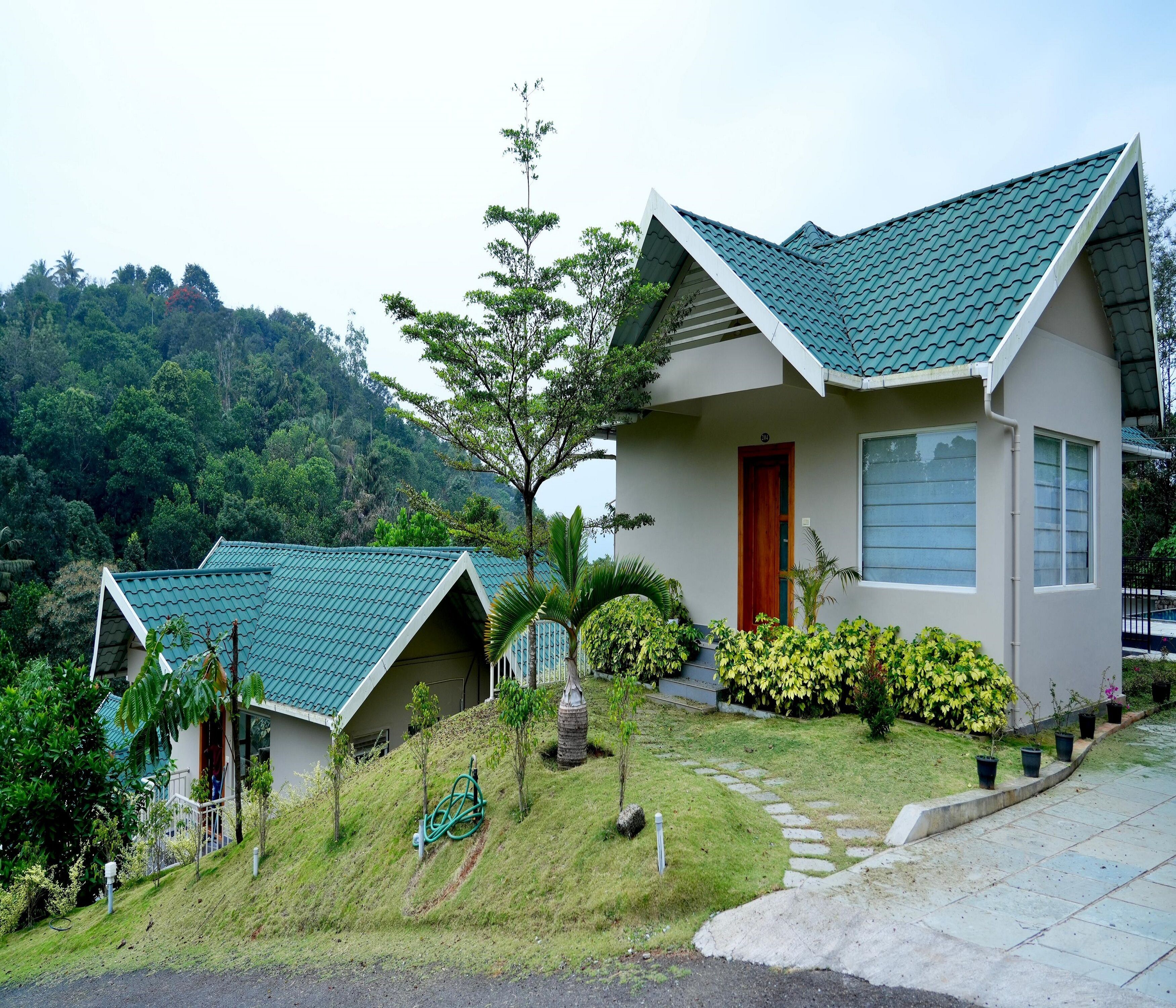 A two-story house with a green roof, surrounded by lush greenery and a hilly landscape.