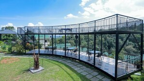 A glass-enclosed outdoor dining area with tables and chairs, surrounded by a metal railing, overlooking a lush landscape. - Aveda Munnar Mountains and Mist (Devikolam)