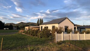 A modern house with a sloped roof, surrounded by a white picket fence, set against a backdrop of hills and a clear sky. - Luxuriate in Lumsden (Lumsden)