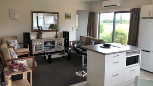 A modern kitchen with a central island, a dining area with a sofa and chairs, a large window with curtains, and a mounted air conditioner. - Luxuriate in Lumsden (Lumsden)