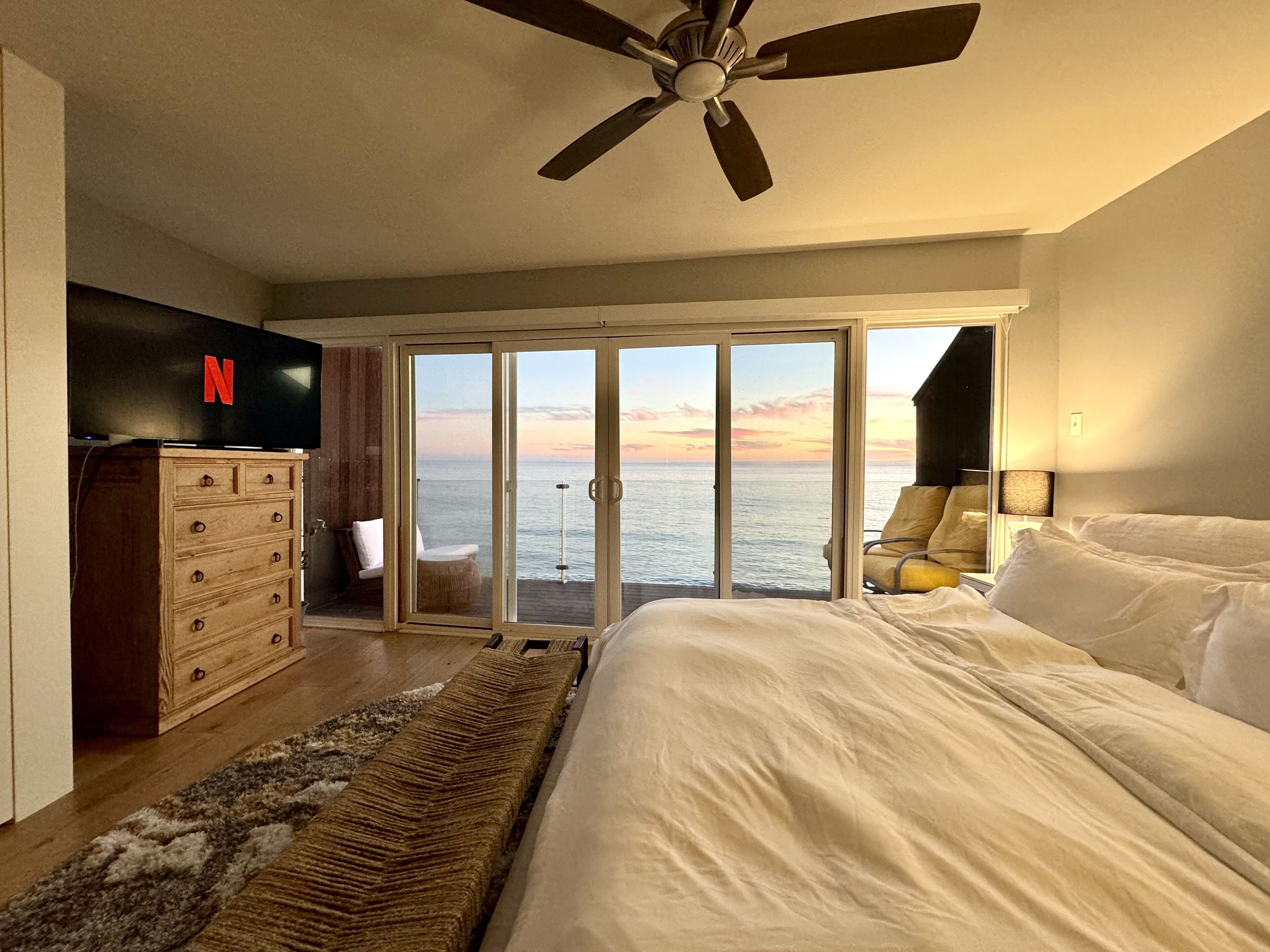 A bedroom with a large bed, a ceiling fan, a wooden dresser, a Netflix logo, a sliding glass door leading to a balcony with a view of the sea, and a nightstand with a lamp.