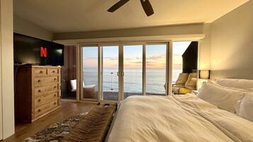 A bedroom with a large bed, a ceiling fan, a wooden dresser, a Netflix logo, a sliding glass door leading to a balcony with a view of the sea, and a nightstand with a lamp.