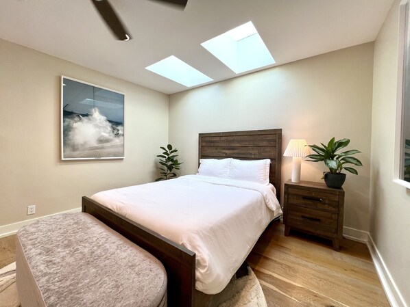 A bedroom with a bed, a nightstand, a lamp, a potted plant, and a window with skylights.