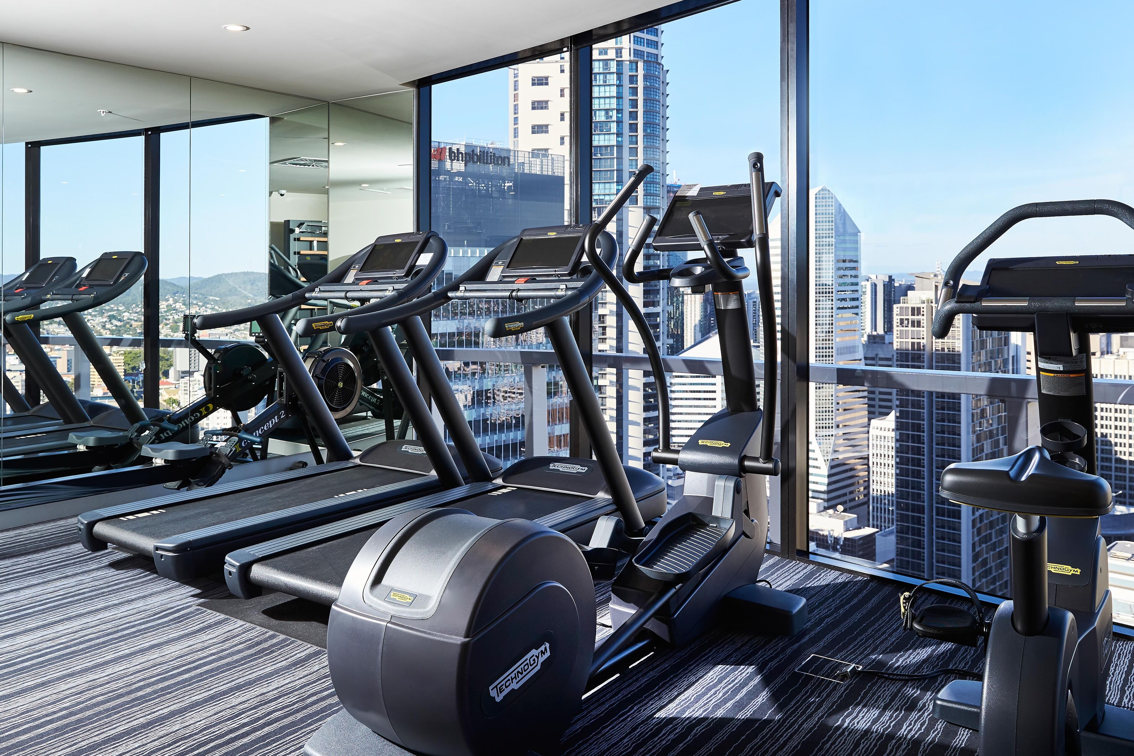 A gym with treadmills and elliptical machines, large windows with a city view, and a carpeted floor.
