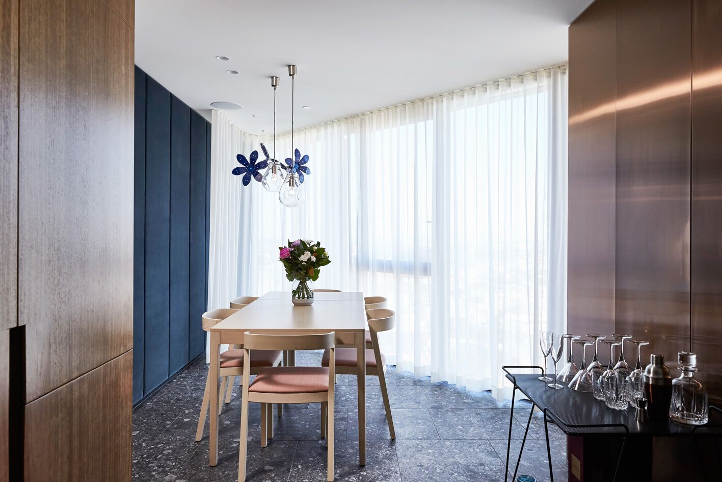 A modern dining area with a wooden table, chairs, and a vase of flowers. There are curtains and a window with blinds.