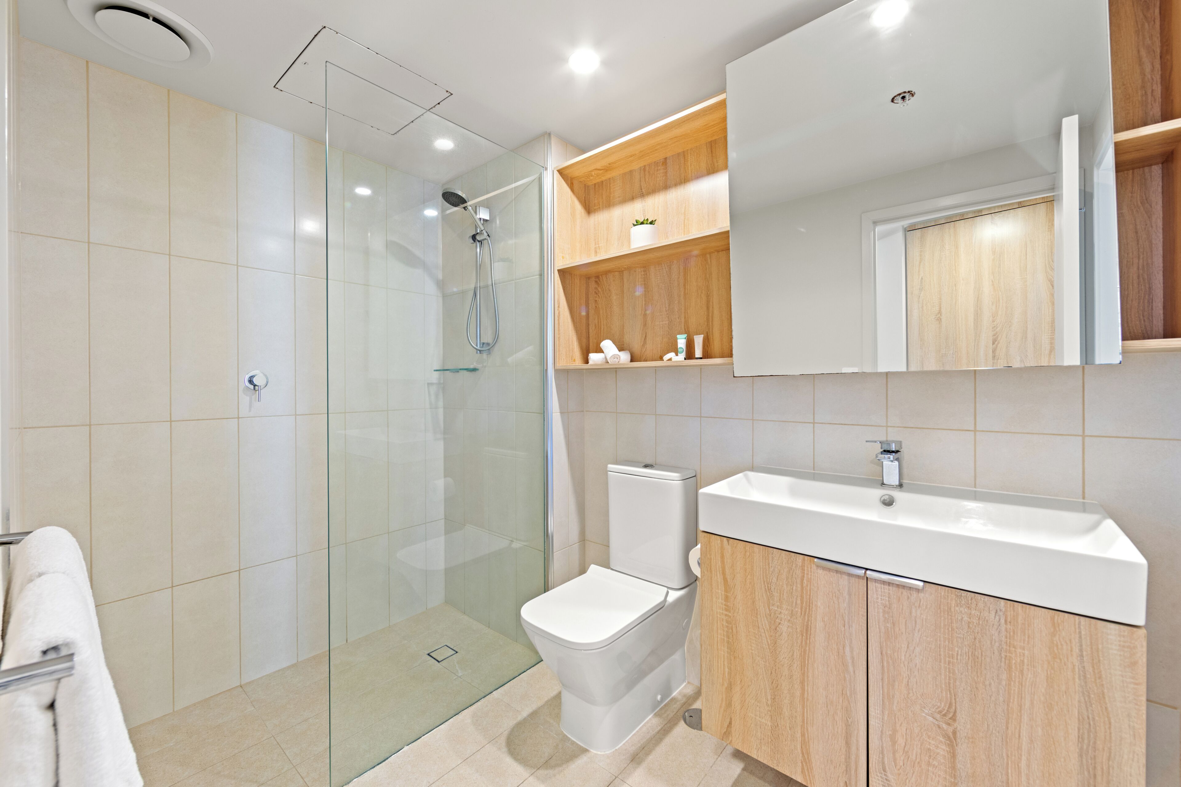Un baño moderno con ducha de vidrio, inodoro blanco, tocador de madera con lavamanos y espejo sobre el tocador.