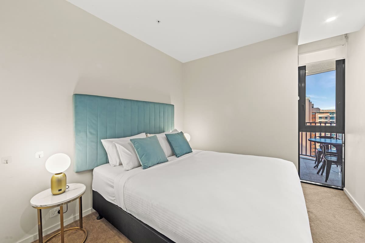 A modern bedroom with a large bed, a bedside table with a lamp, and a view of the outside balcony.