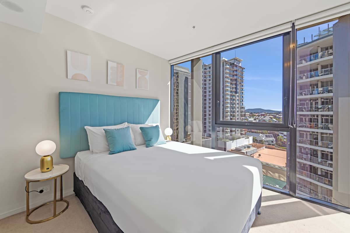 A bedroom with a large bed, a nightstand, a window with a view of buildings, and a ceiling light.