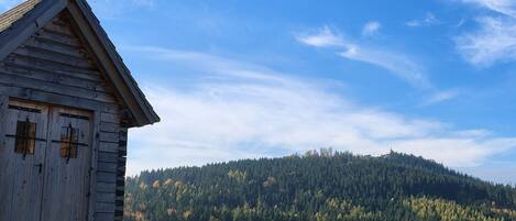 A wooden church with a cross on top, set against a backdrop of a forested hill and clear sky.