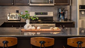 A modern kitchen with dark cabinets, stainless steel appliances, a central island with chairs, and a decorative plant. - Agave Zen House w/ Hot Tub & BBQ Grill! (Porterville)