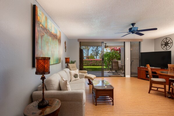 A spacious living room with a sofa, coffee table, and dining area. There is a ceiling fan, a TV, and a painting on the wall.