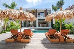 A poolside area with wooden lounge chairs and thatched umbrellas, a modern house in the background, and lush greenery. - Villa Tortuga (Canggu)