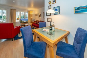 A modern living room with a glass-top dining table, blue upholstered chairs, a red sofa, and a view of the outdoors through large windows.
