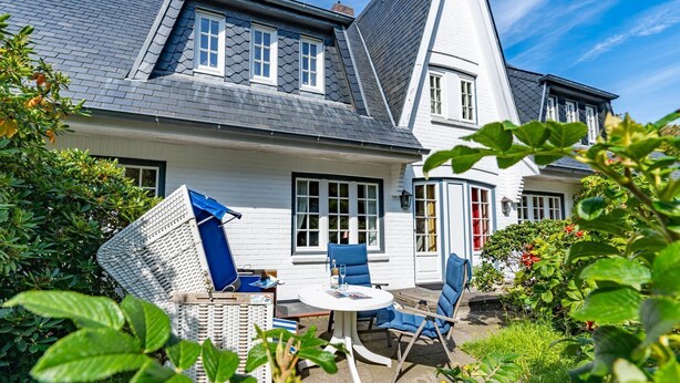 Une maison avec un toit en pente, des murs extérieurs blancs et un petit patio comprenant une table ronde et des chaises bleues.