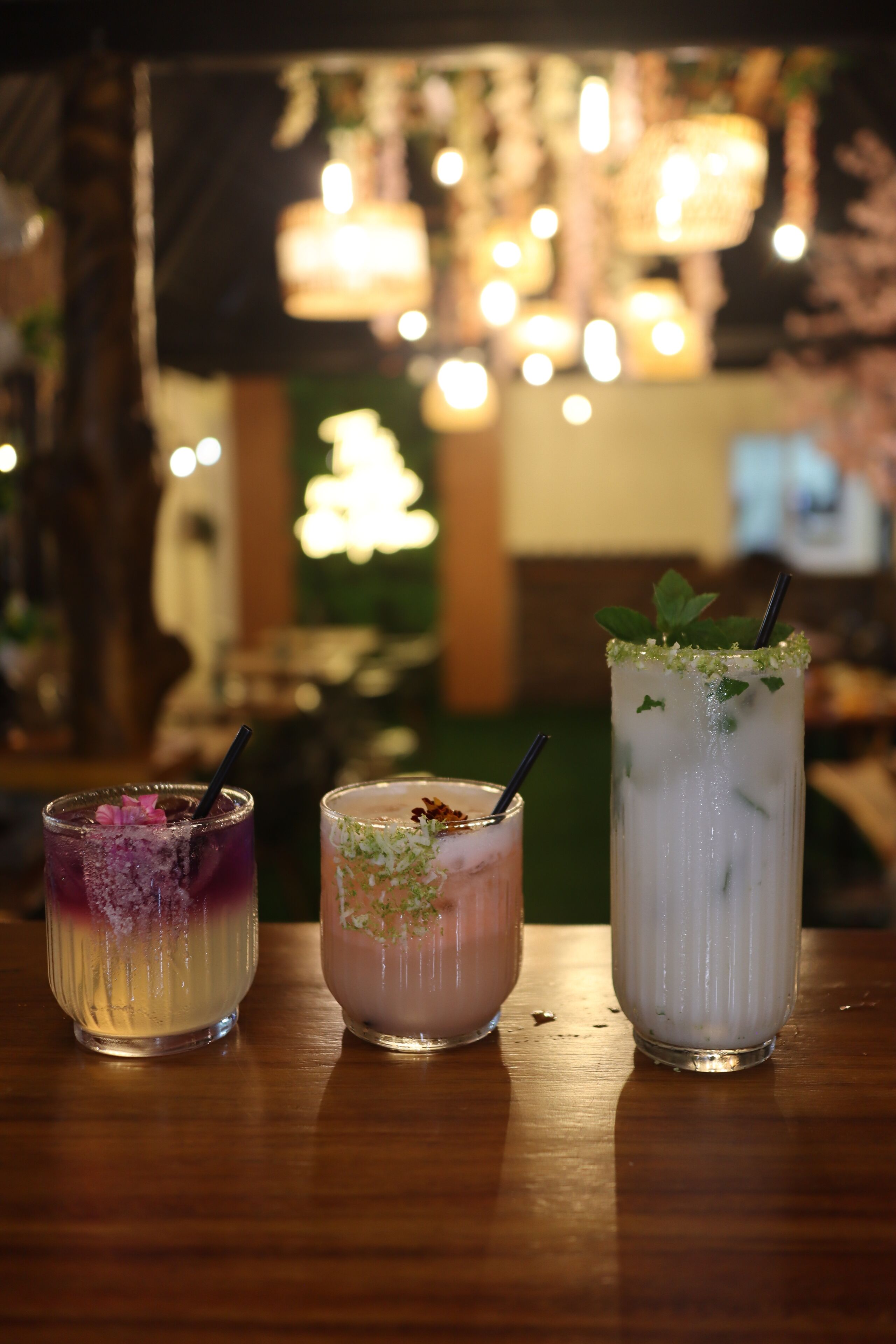 Three drinks on a wooden table with a blurred background of hanging lights.
