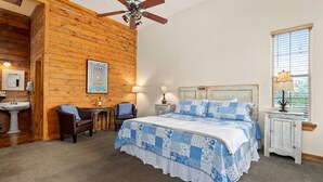 Un dormitorio con pared de madera, una cama con ropa de cama azul y blanca, un ventilador de techo, una ventana con persianas y una pequeña mesa con lámpara.